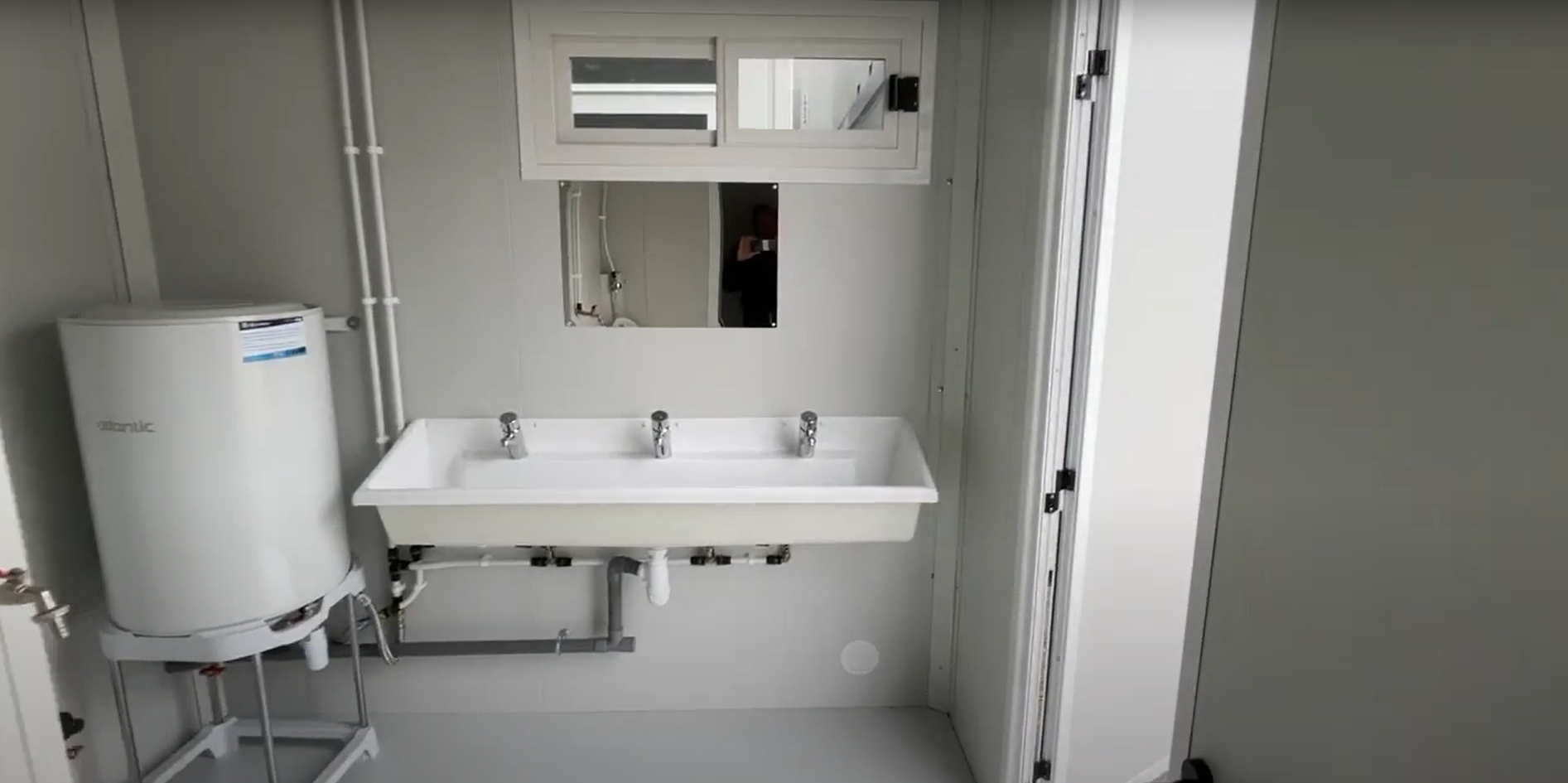 L'intérieur d'un bungalow de chantier sanitaire, on y voit en face un lavabo avec 3 robinets avec au-dessus un miroir et une petite fenêtre. Sur la gauche, en bas, un chauffe eau de 100L. A droite se trouve la porte de sortie.