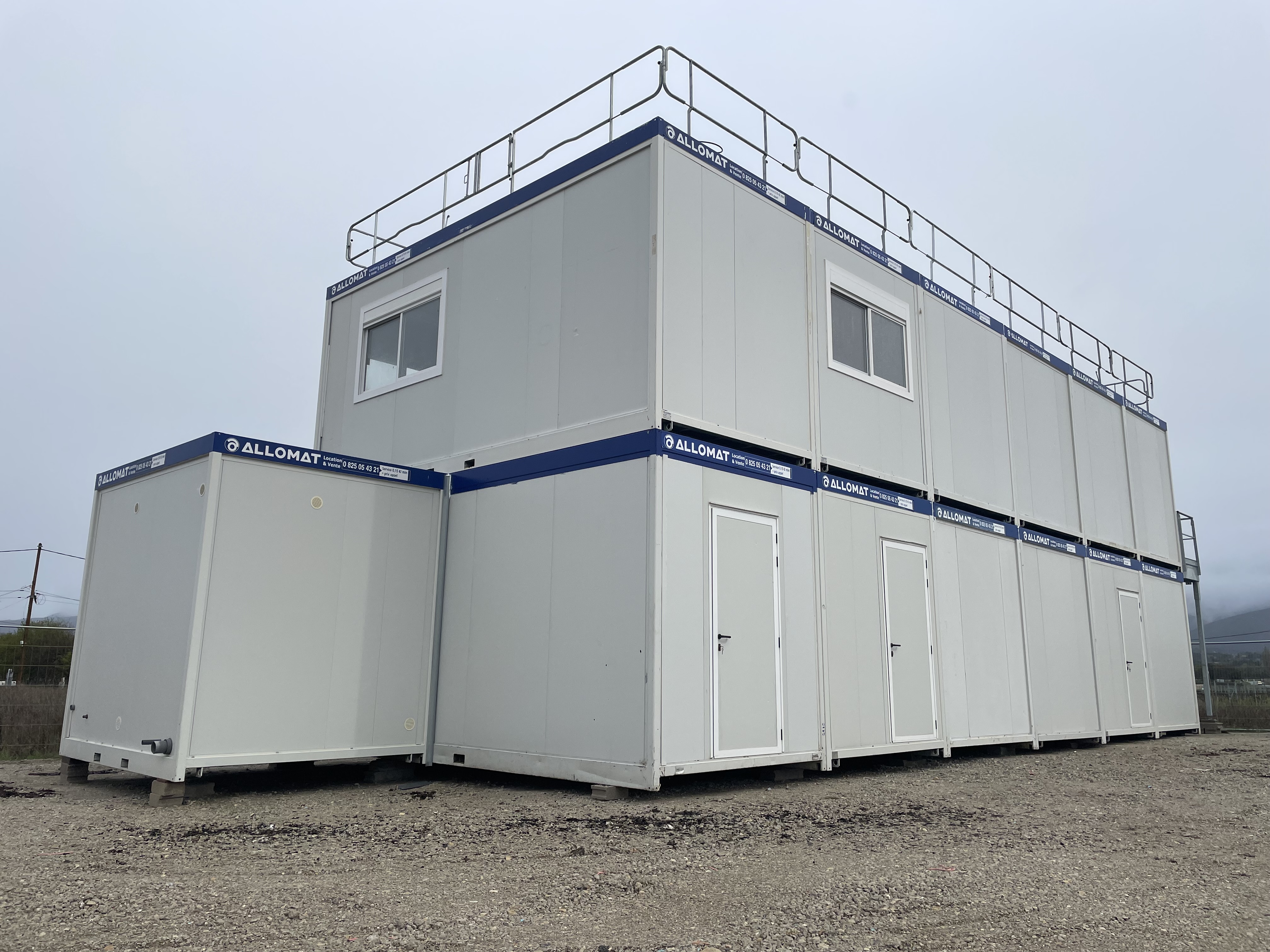 Photo d'une base vie de chantier, des modules gris à bords bleus, avec des gardes corps sous un ciel couvert de nuages