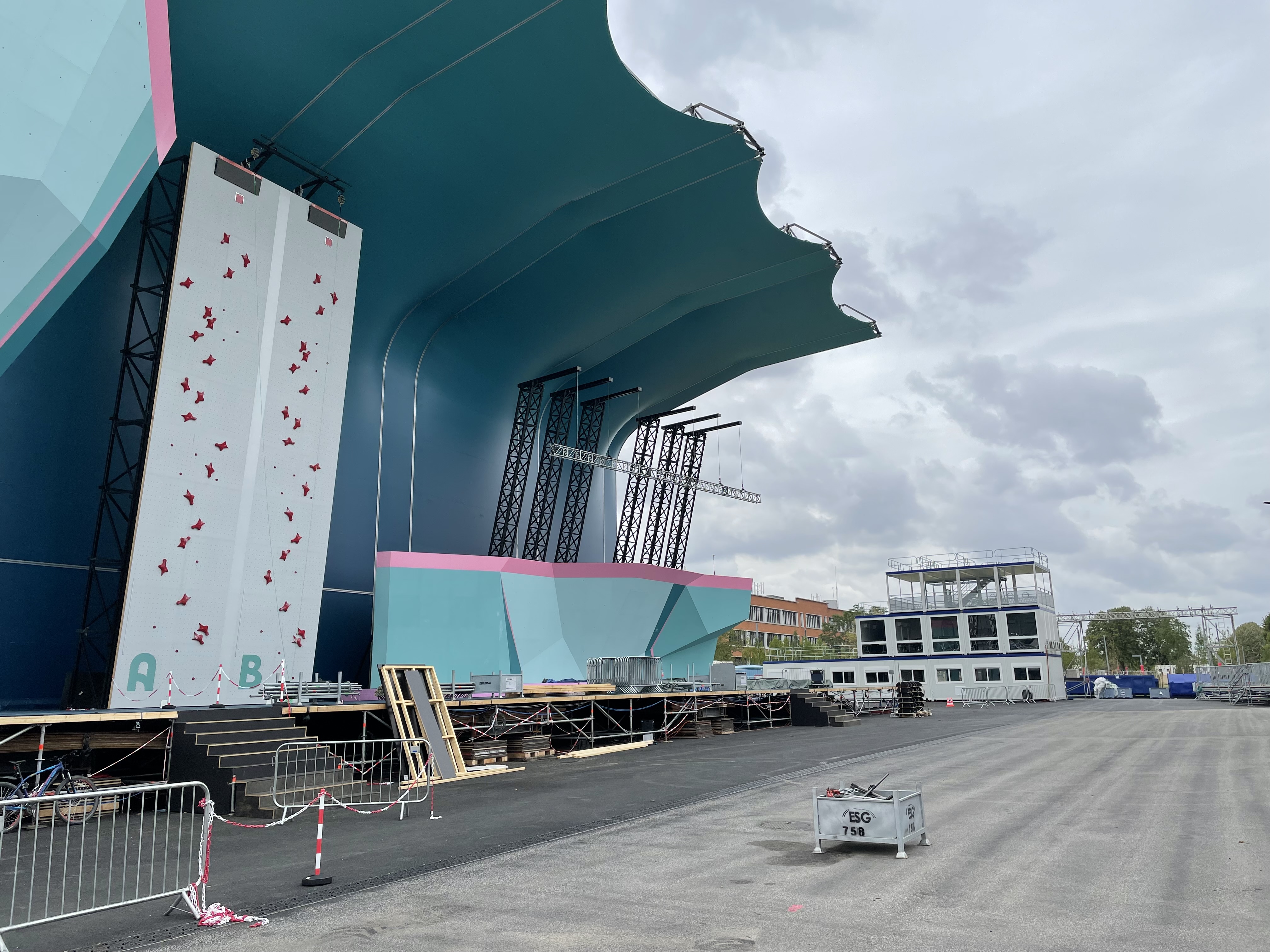 Le mur d'escalade des JO Paris 2024, un mur blanc grisé avec des prises de toutes les couleurs. Il est sous un abri vert bleu océan. Au fond on voit la régie temporaire de l'événement, composée de modules ALLOMAT, sur 2 étages avec une terrasse sur le toit.