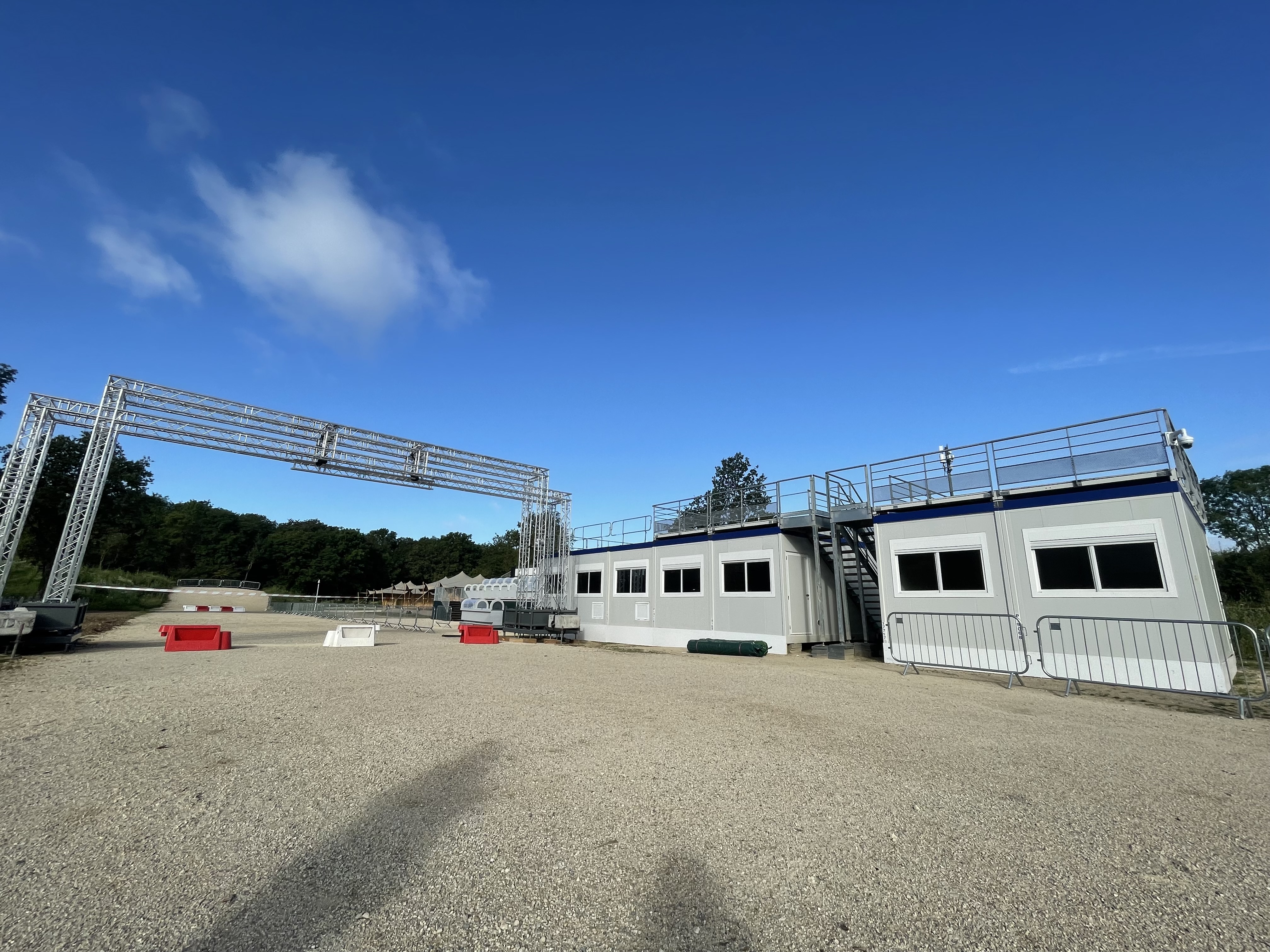 La ligne d'arrivée de la course de VTT des JO. On peut y voir les modules temporaires ALLOMAT abritant les commissaires de courses alignés sur le côté de la piste en gravier. Il y a 2 escaliers permettant d'accéder à la terrasse présent sur le toit des modules. Sur la gauche, la piste se poursuit sous l'arche métallique, de la rubalise interdit le passage, l'événement n'ayant pas encore eu lieu. Il y a aussi des plots blancs et rouges au milieu de la piste interdisant de la pratiquer. Au fon se trouve une bosse avec les anneaux olympiques dessinés. Il y a de grands arbres feuillus au fond.