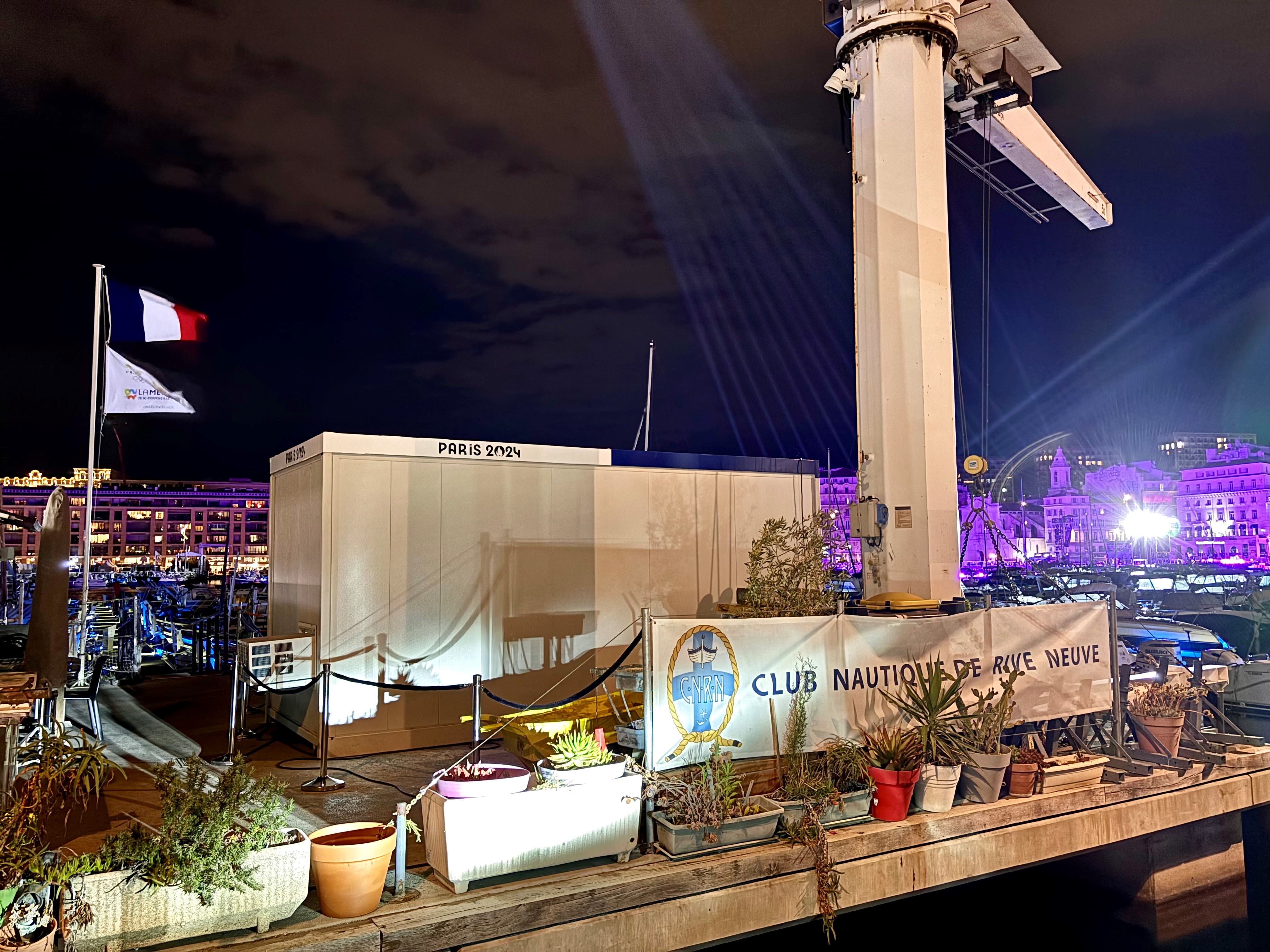 Un module de construction temporaire de nuit sur le Vieux-Port, aux couleurs de Paris 2024. Il est éclairé par une lumière violette en arrière plan de la scène du concert