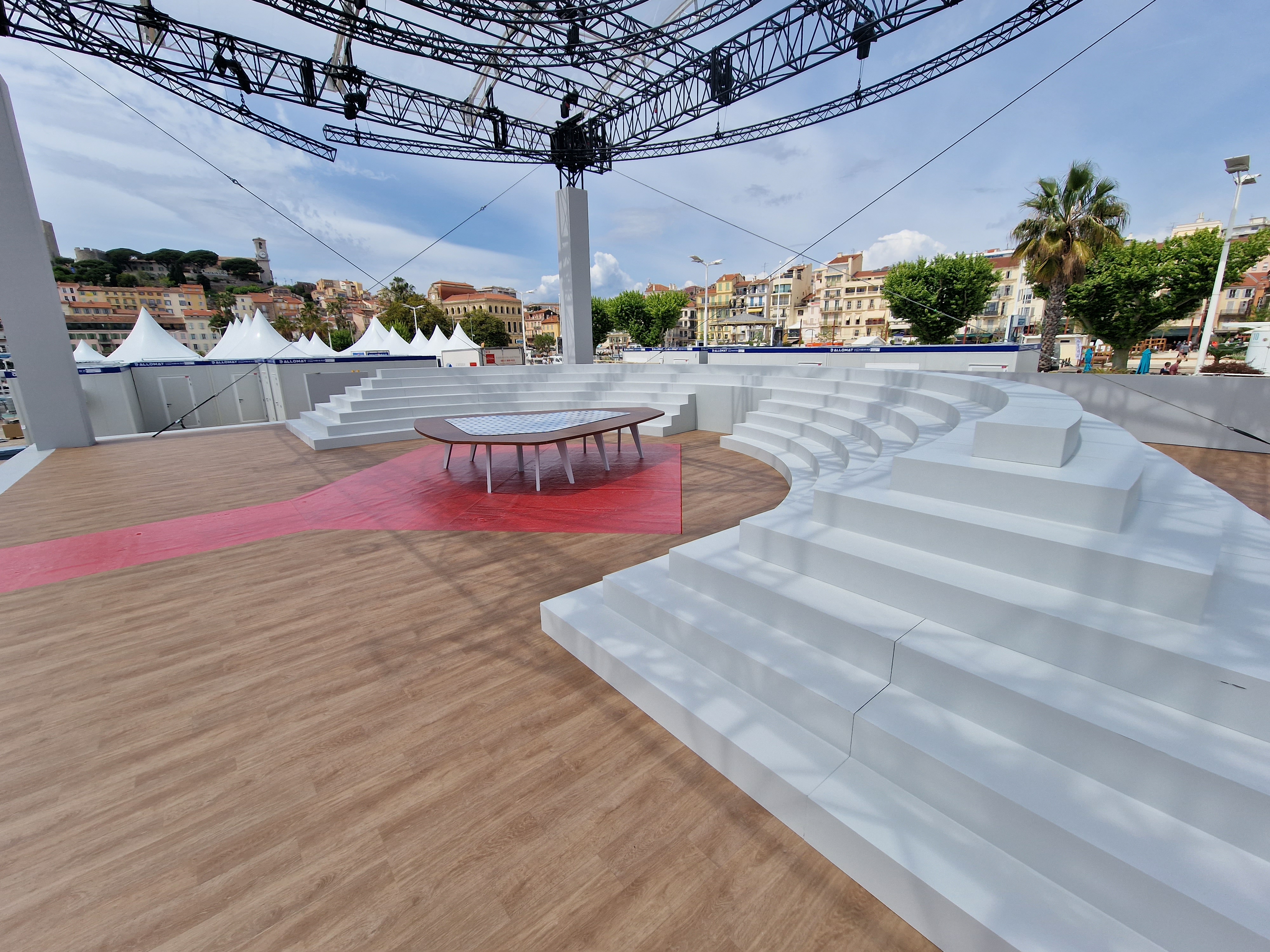 Le plateau du festival de Cannes de 2023. On voit un petit amphithéâtre blanc entourant une grande table triangulaire posée sur un tapis rouge qui part vers la gauche de la photo, sur un sol en parquet. Au second plan, sur la gauche, on voit des chapiteaux blanc posés sur une construction modulaire Allomat composée de plusieurs modules avec la ville au fond. Idem sur la droite, au second plan on voit le haut de modules de la gamme Domino d'Allomat avec la ville au fond