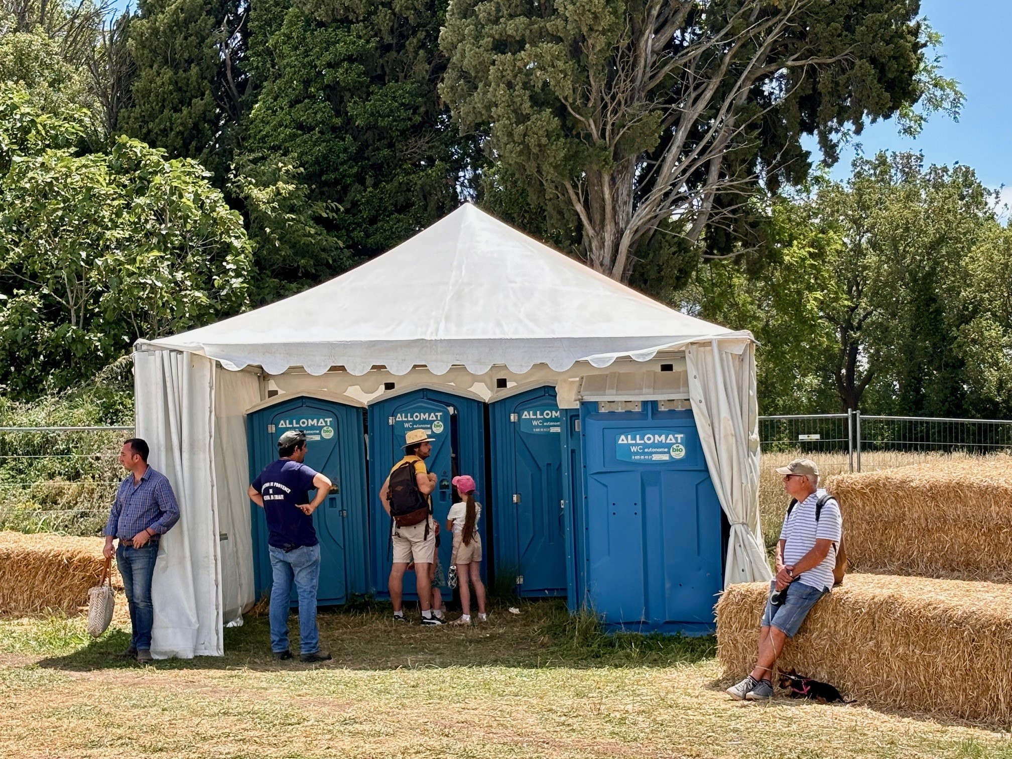 La photo montre une grande tente blanche où sont installés 5 toilettes autonomes allomat bleus foncés. Il y a autour des bottes de foin. 5 personnes attendent. La plus à gauche est un homme blanc aux cheveux brun gris, avec une chemise à carreaux et un jean bleus. Il porte aussi un sac blanc et il est appuyé sur le pied gauche de la tente. Une deuxième personne attend dehors, les mains sur les hanches. Il porte une casquette verte, un t-shirt bleu avec une inscription blanche et un jean bleu. Au milieu, devant les WC autonomes Allomat, un père et sa fille attendent devant la porte. Le père porte un chapeau blanc avec une bande noire, un chemise jaune et un sac à dos noir. Il a aussi un bermudas blanc et des baskets blanche et bleues. sa fille porte une casquette rose, un t-shirt blanc, des cheveux longs tirés en queue de cheval à travers l'anneau de serrage de la casquette et un short beige. La dernière personne est assise en dehors de la tente, à droite, sur une botte de foins. C'est un homme un peu âgé, qui porte une casquette beige, un t-shirt rayé type marinière bleue et blanche, un short en jean et des baskets en toile. Il tient en laisse un petit chien noire et jaune sur le poitrail.